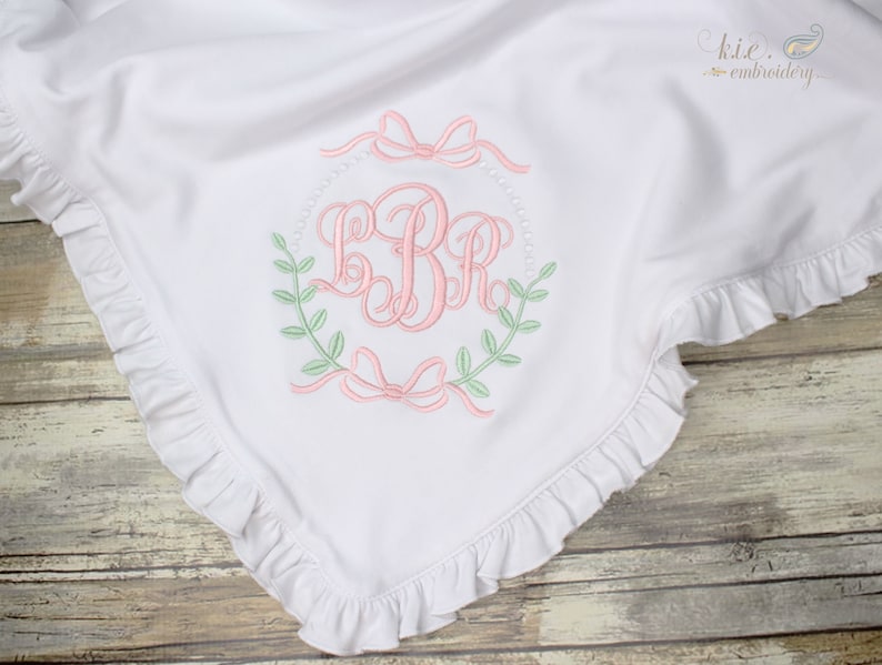 Op de afbeelding: Wit babydekentje met een gerimpelde rand en een geborduurd monogram in het midden. Het monogram is omringd door een krans van groene bladeren en roze strikken.