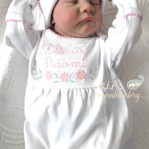 May include: A white baby gown and matching cap with pink trim. The gown features the embroidered names "Olivia Naomi" above floral designs. The baby is wearing the gown and cap.