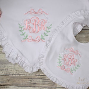 Op de afbeelding: Een wit babydekentje en een slabbetje met roze en groene borduursels. Zowel het dekentje als het slabbetje zijn afgewerkt met ruches. De borduursels bevatten een monogram met de initialen "LBR" omringd door een krans en strikken.