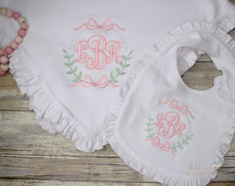 Op de afbeelding: Een wit babydekentje en een slabbetje met roze en groene borduursels. Zowel het dekentje als het slabbetje zijn afgewerkt met ruches. De borduursels bevatten een monogram met de initialen "LBR" omringd door een krans en strikken.