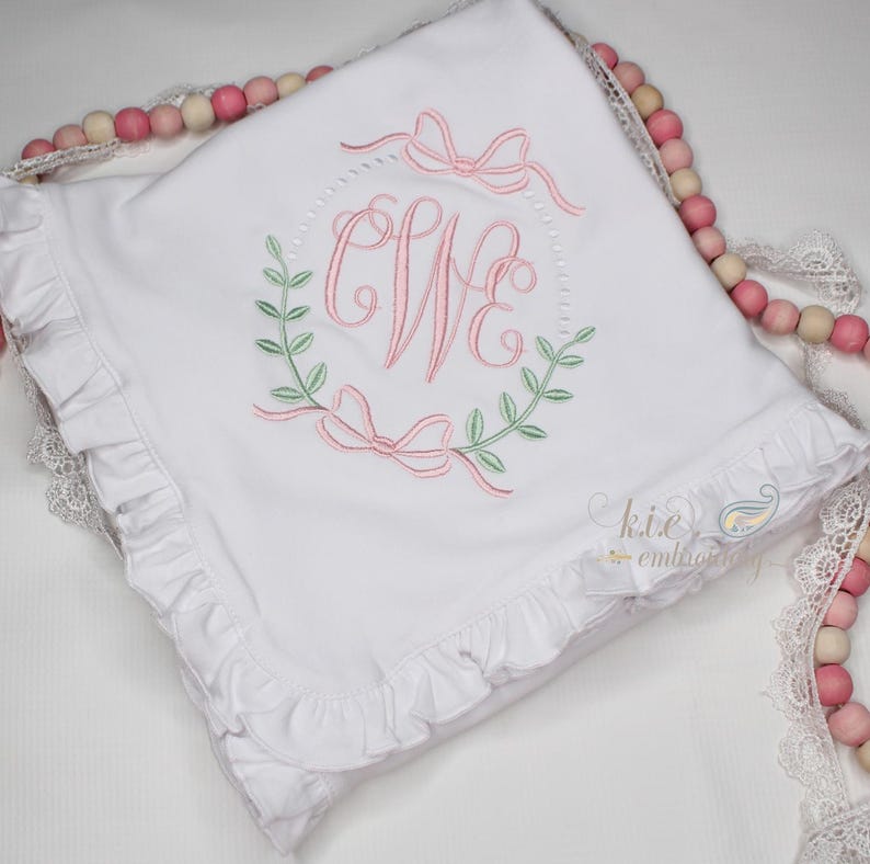 Op de afbeelding: Een wit babydekentje met een gerimpelde rand en een geborduurd monogram in het midden. Het monogram is omgeven door een krans van bladeren en strikken. Het dekentje is gemaakt van zacht katoen.