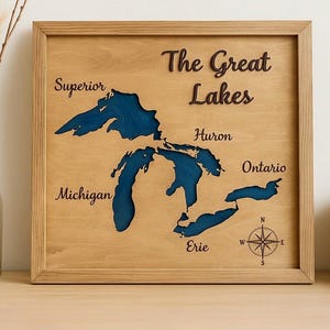 May include: Wooden framed wall art featuring a map of the Great Lakes. The lakes are depicted in blue against a light brown wood background. The names of the lakes are labeled in a cursive font. A compass rose is in the lower right corner.