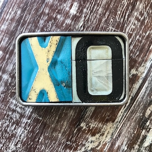 May include: A silver belt buckle with a blue, white, and black design. The buckle features the letters "XO" in a vintage style.
