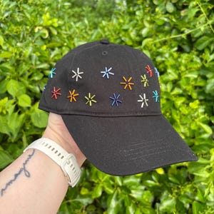 May include: A black baseball cap featuring a pattern of small, multi-colored star-shaped embroidered accents in shades of red, orange, yellow, blue, and white scattered across the front and side panels of the crown.