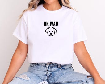May include: White t-shirt with the text "OK WAU" above a simple dog face graphic in black. The shirt is made of a soft material and is suitable for casual wear. The model is wearing blue jeans.