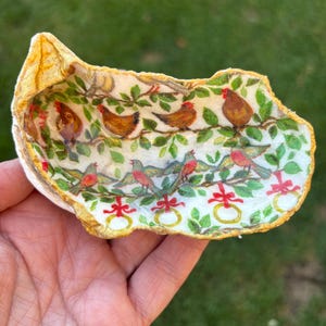 May include: A decorative, hand-painted oyster shell. The interior features a design of brown chickens and red birds perched on green leafy branches. The shell's edge is trimmed with gold paint, and the background is a blurred green.