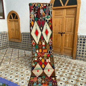 Op de afbeelding: Een lang, smal, kleurrijk tapijt met een geometrisch patroon. Het tapijt is bruin met witte, rode, gele, blauwe en groene accenten. Het tapijt hangt aan een muur in een kamer met betegelde vloer.