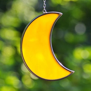 Könnte beinhalten: Ein gelber Glas-Halbmond-Sonnenfänger, der an einer silbernen Kette hängt. Der Sonnenfänger hängt vor einem verschwommenen grünen Hintergrund.