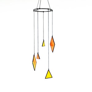 Könnte beinhalten: Hängendes Mobile mit einem schwarzen Metallring und Schnüren, an denen geometrische Formen befestigt sind. Die Formen sind orange und gelbe Rauten und Dreiecke mit schwarzen Umrissen. Der Hintergrund ist weiß.