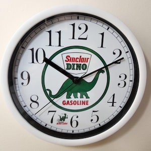 May include: A round wall clock with a white frame and a white face featuring black numerals. The center displays a green Sinclair Dino logo with the text Sinclair DINO GASOLINE and a small Dino Lube graphic near the bottom. Black clock hands.
