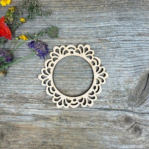 Könnte beinhalten: Ein lasergeschnittener dekorativer Holzring mit gewelltem Rand. Der naturfarbene Holzring ist von getrockneten Blumen umgeben und liegt auf einer verwitterten Holzoberfläche.