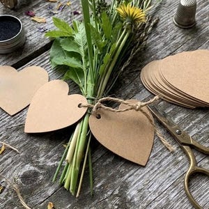 Può includere: Un mazzetto di erba verde e un fiore di tarassaco giallo legati con dello spago a due etichette di carta marrone a forma di cuore. Vicino si trovano un ditale in metallo, una lente d'ingrandimento, un timbro in legno, cera nera e forbici.
