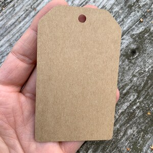 Pode incluir: Uma etiqueta de presente de papel castanho com um furo perfurado no topo. A etiqueta est&aacute; em branco e pronta para ser escrita.