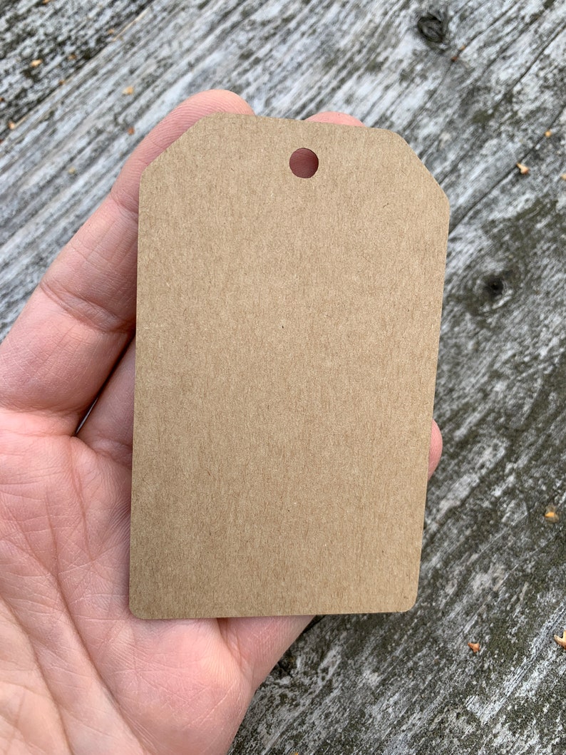 Pode incluir: Uma etiqueta de presente de papel castanho com um furo perfurado no topo. A etiqueta est&aacute; em branco e pronta para ser escrita.