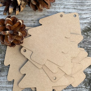 K&ouml;nnte beinhalten: Ein Stapel brauner Papiertannenbaum-Geschenk-Etiketten mit oben gelochten L&ouml;chern. Die Etiketten liegen auf einer Holzoberfl&auml;che mit Tannenzapfen.