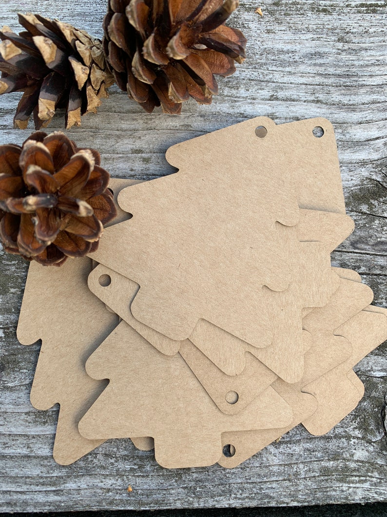K&ouml;nnte beinhalten: Ein Stapel brauner Papiertannenbaum-Geschenk-Etiketten mit oben gelochten L&ouml;chern. Die Etiketten liegen auf einer Holzoberfl&auml;che mit Tannenzapfen.