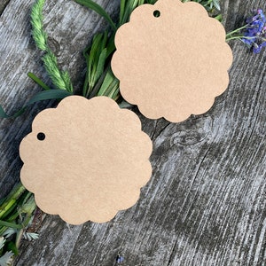 May include: Two brown paper gift tags with scalloped edges and a hole punched at the top. The tags are lying on a wooden surface with green plants and flowers.