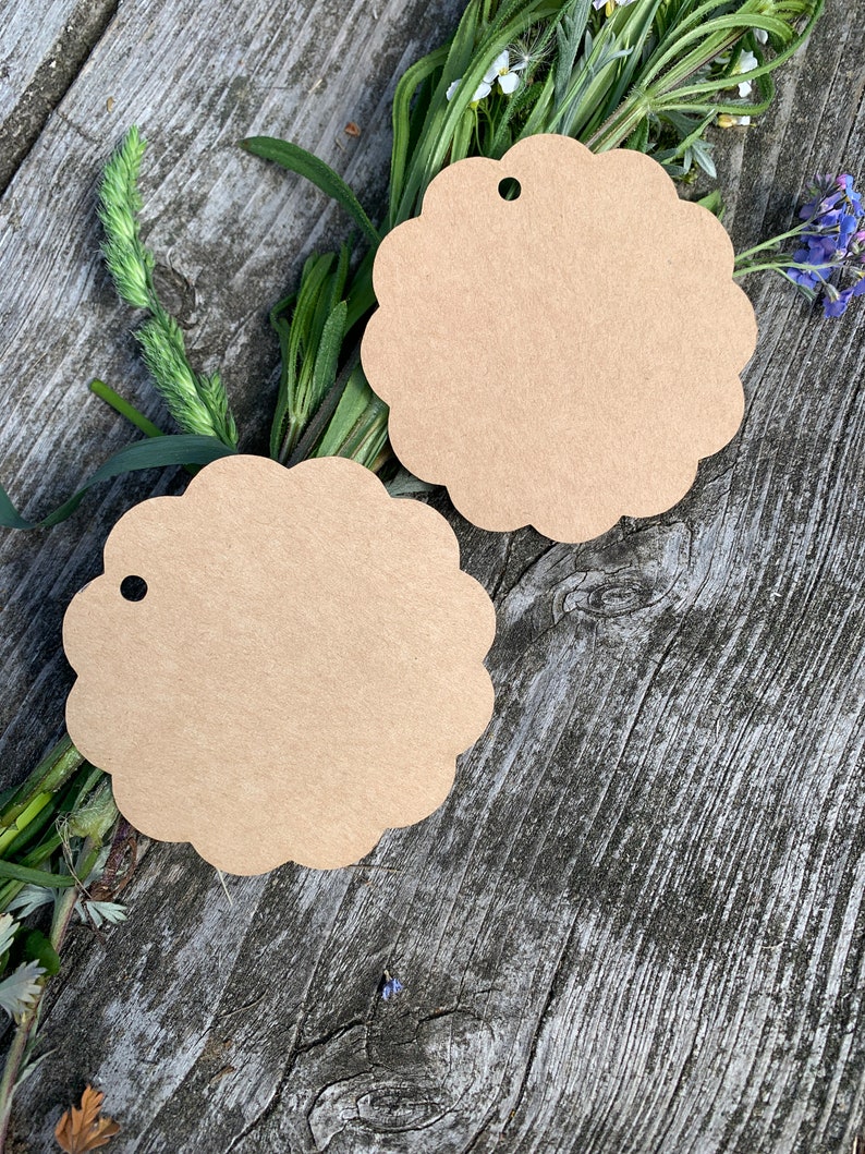 May include: Two brown paper gift tags with scalloped edges and a hole punched at the top. The tags are lying on a wooden surface with green plants and flowers.