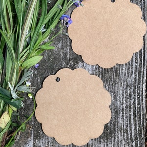 May include: Two brown paper gift tags with scalloped edges and a hole punched in the top. The tags are on a weathered wooden surface with blue flowers.