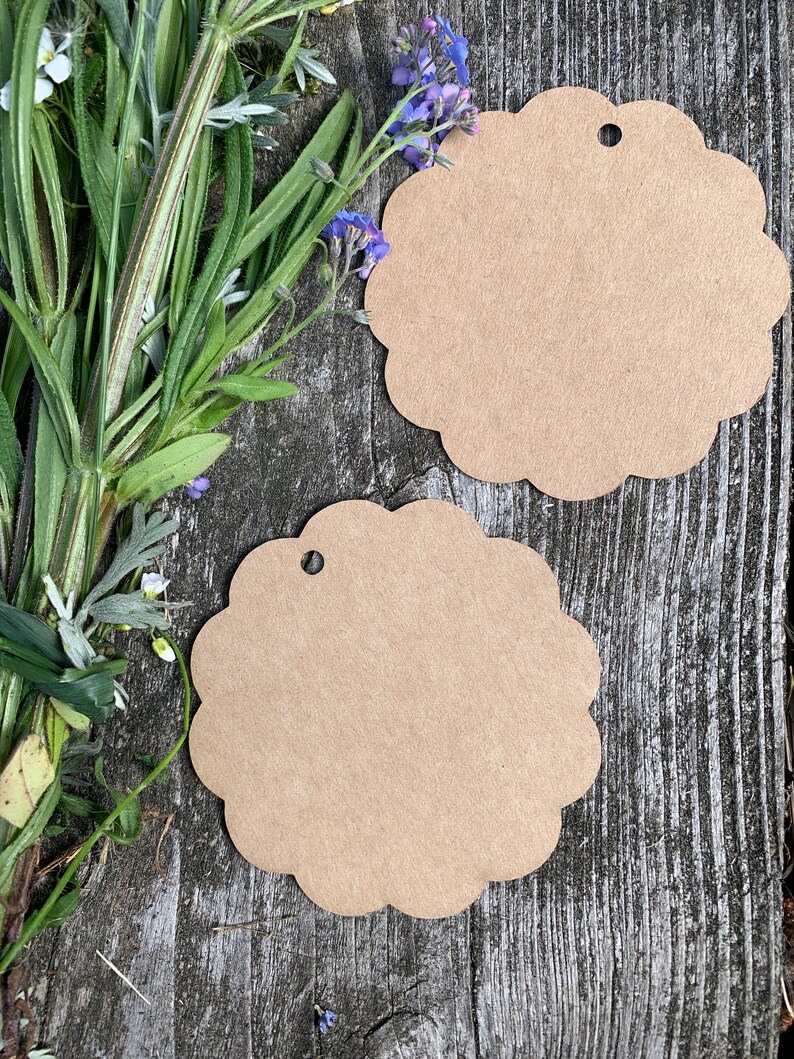 May include: Two brown paper gift tags with scalloped edges and a hole punched in the top. The tags are on a weathered wooden surface with blue flowers.