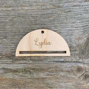 Puede incluir: Un adorno de madera en forma de semicírculo con el nombre "Lydia" grabado en él. El adorno tiene un agujero en la parte superior para colgarlo.