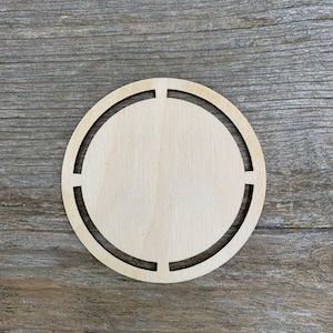 Puede incluir: Un recorte redondo de madera con un diseño circular interior decorativo. El recorte está sin terminar y listo para ser pintado o teñido.
