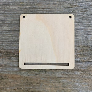 Può includere: Un cartello di legno vuoto con due fori nella parte superiore e una fessura nella parte inferiore. Il cartello è in legno marrone chiaro e ha una superficie liscia.