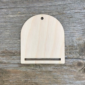 May include: A wooden cutout in the shape of an arch with a small hole at the top and a horizontal slot at the bottom. The cutout is made of light-coloured wood.