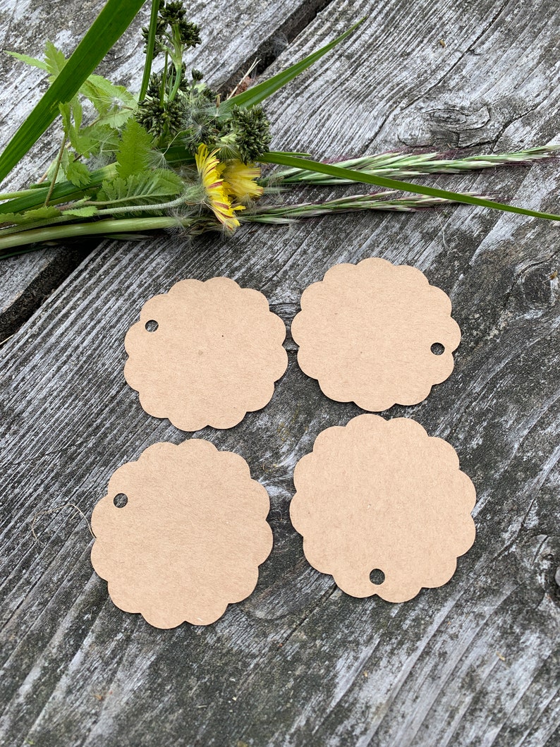 May include: Four brown paper gift tags with scalloped edges and a hole punched in the top. The tags are arranged in a square shape on a wooden surface.