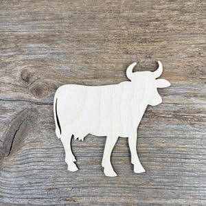 Puede incluir: Un recorte de madera plana y de color claro con la forma de una vaca vista de perfil. La silueta presenta una cabeza distintiva con dos cuernos curvados, un cuerpo largo, cuatro patas y una cola. La superficie muestra una veta natural.