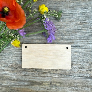 May include: A rectangular, unfinished wooden tag with two pre-drilled holes, resting on a weathered wooden surface. A vibrant arrangement of wildflowers, including a large orange poppy, yellow buttercups, and purple scabious, adds a touch of color.