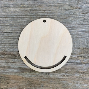 Puede incluir: Un círculo de madera con una forma de sonrisa recortada. El círculo tiene un agujero en la parte superior para colgar.