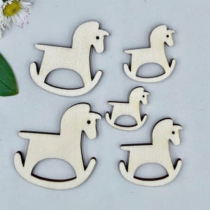 May include: Five wooden rocking horse cutouts in varying sizes. The horses are light-coloured with dark brown outlines, and are arranged on a light grey surface. A daisy and green leaf are in the upper left corner.