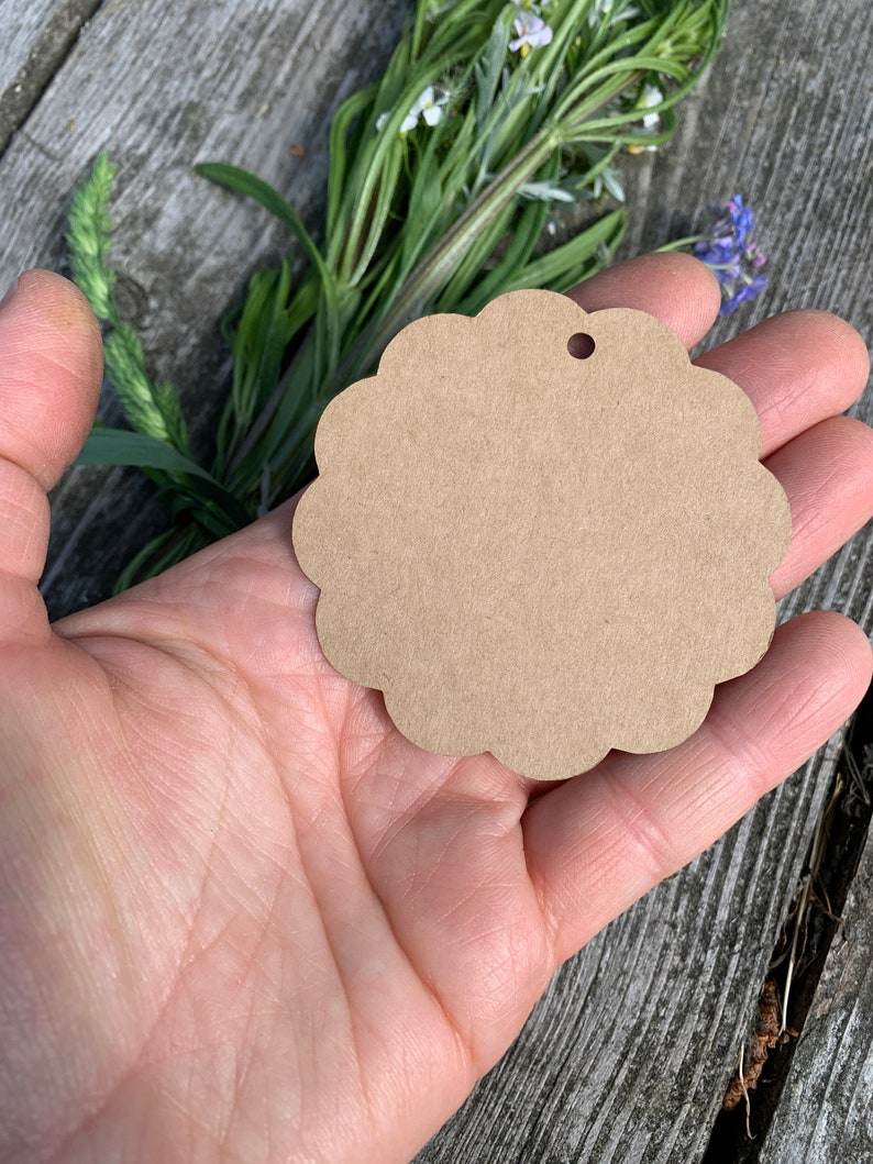 May include: A brown, scalloped, circular gift tag with a hole punched in the top. The tag is held in a person's hand.