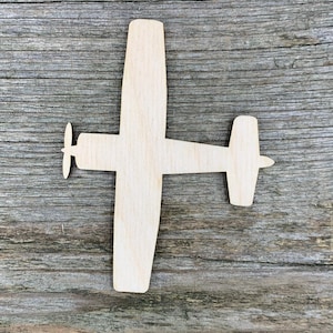 Pode incluir: Um recorte de madeira plano e de cor clara em forma de um pequeno avião. A silhueta apresenta uma fuselagem central, duas asas longas que se estendem pelos lados, uma hélice na parte frontal e um estabilizador horizontal com cauda.