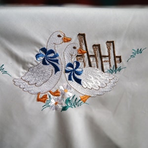 Könnte beinhalten: Weiße bestickte Tischdecke mit zwei weißen Gänsen mit blauen Schleifen, einem braunen Zaun und grünem Laub.