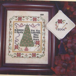 May include: A framed cross-stitch sampler with a Christmas tree design. The sampler is titled "Rejoice O Earth" and includes the text "For the Savior has been born". The sampler is in a brown frame with a white mat. The sampler is surrounded by red fall leaves.