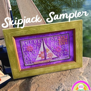 May include: A framed cross-stitch sampler with a purple background and a sailboat design. The sampler includes the alphabet, numbers, and a variety of star and geometric patterns. The frame is gold with a brown inner edge. The title "Skipjack Sampler" is written in white cursive letters above the frame.
