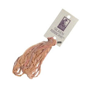 Peut inclure: Un lot de rubans de soie aux tons pêche et rose, attachés à une étiquette crème. L'étiquette porte l'inscription "Silken Ribbons" et un logo. Les rubans sont lâchement bouclés, créant un aspect texturé.