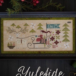 May include: A framed cross-stitch sampler featuring a white deer pulling a sleigh with a figure in red. The sampler is titled "Yuleside Delivery" and includes the text "Dec. 25".