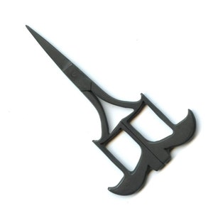 May include: Black metal scissors with a unique, ornate design. The blades are closed and the handles are shaped like a stylised letter 'M'.