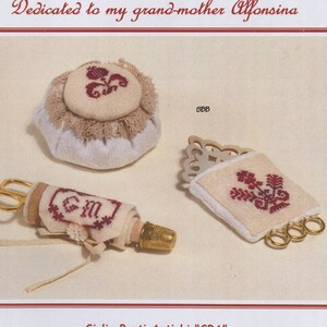 May include: A vintage-style sewing kit with a pincushion, needle threader, and scissors. The pincushion is white with a red embroidered flower design. The needle threader is a small wooden spool with a red embroidered design. The scissors are gold with a red embroidered design. The kit is dedicated to Alfonsina.