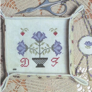Può includere: Uno schema di ricamo a punto croce per un vassoio floreale e un portachiavi per forbici. Il disegno presenta una pianta in vaso con tre fiori viola e foglie verdi. Il testo "The Mindful Needle Designs" è in un cerchio nella parte superiore dell'immagine. Il testo "Designed by Dawn Friske MN-022" è nella parte inferiore dell'immagine.
