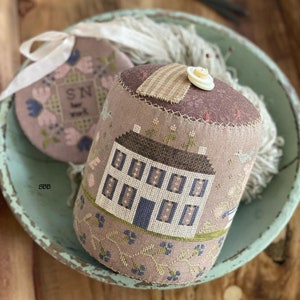 May include: A brown and beige pincushion with a white house design and blue trim. The pincushion is decorated with small flowers and birds. The pincushion is sitting on a blue and white bowl with a white button on top.