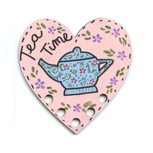 May include: A pink heart-shaped wooden sign with a blue teapot and the words "Tea Time" written in black. The teapot has a floral pattern and is surrounded by small pink and purple flowers.