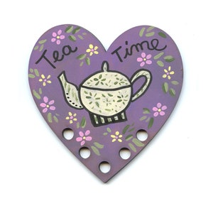 May include: Purple heart-shaped wooden tea cozy with a white teapot and floral design. The text "Tea Time" is written on the cozy.