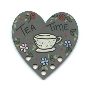 Può includere: Un copri-teiera in legno grigio a forma di cuore con una tazza da tè dipinta e le parole "Tea Time". Il copri-teiera ha dei fori intorno al bordo per cucire.
