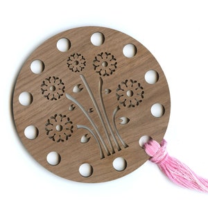 May include: A wooden embroidery floss organizer with a floral design. The round organizer is made of light brown wood and features cut-out flower shapes and holes for holding floss. A bundle of pink embroidery floss is attached.