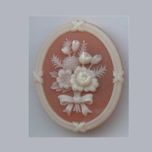 May include: A pink cameo brooch with a white floral design. The cameo is oval shaped and has a white border. The flowers are carved in relief and are arranged in a bouquet.