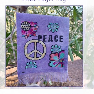 May include: A purple cross stitch pattern of a peace sign with the word "PEACE" above it. The peace sign is surrounded by colorful flowers. The pattern is on a purple fabric with a fringe border.